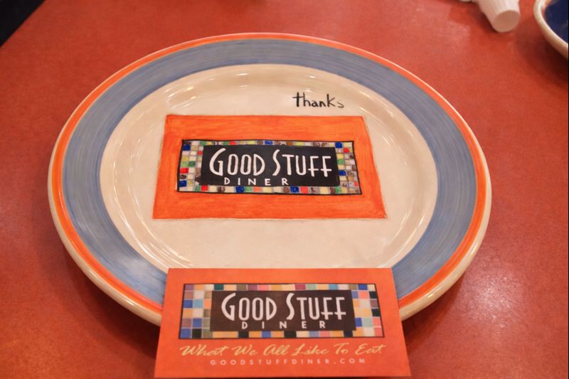 Assiette peinte du logo Good Stuff Diner posée sur une table, avec une carte du restaurant au premier plan.
