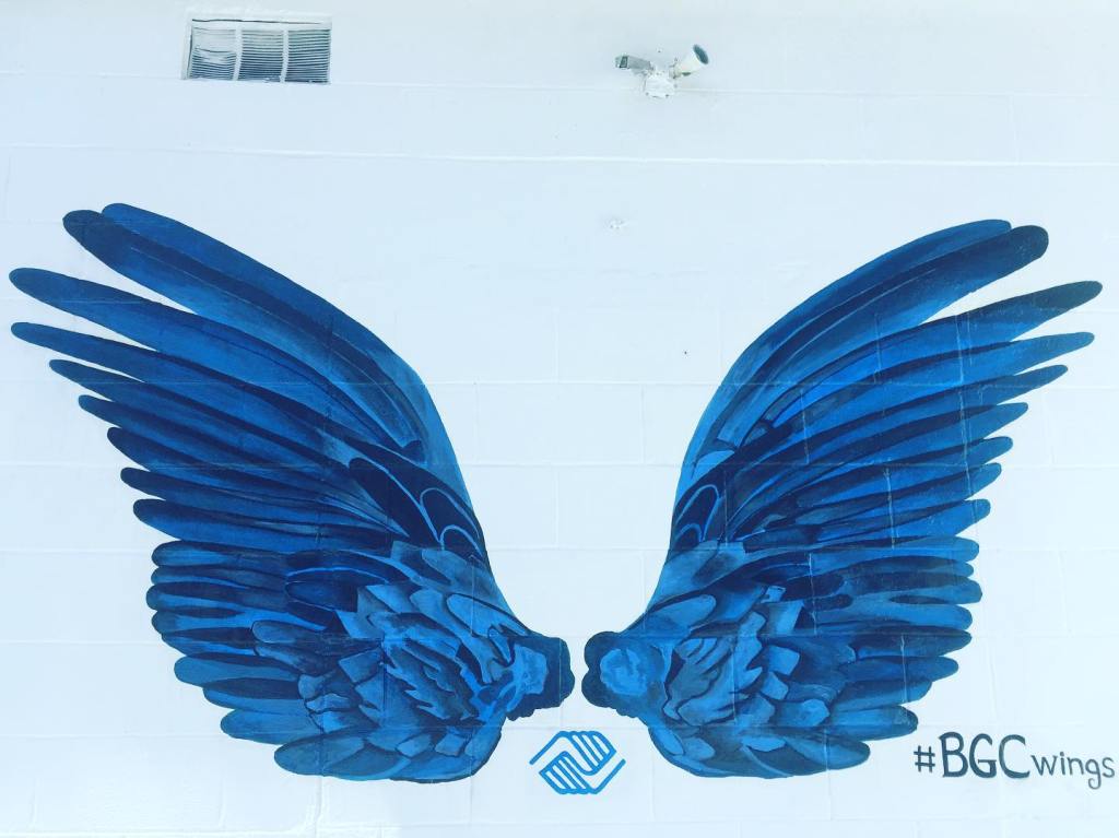 Deux grandes ailes bleues peintes sur un mur blanc avec le logo du Boys & Girls Club au centre.
