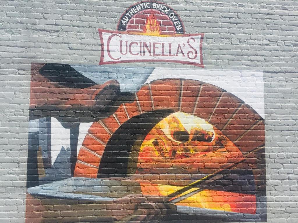 Fresque sur mur de briques représentant un four à pizza en flammes sous le logo Cucinella’s.
