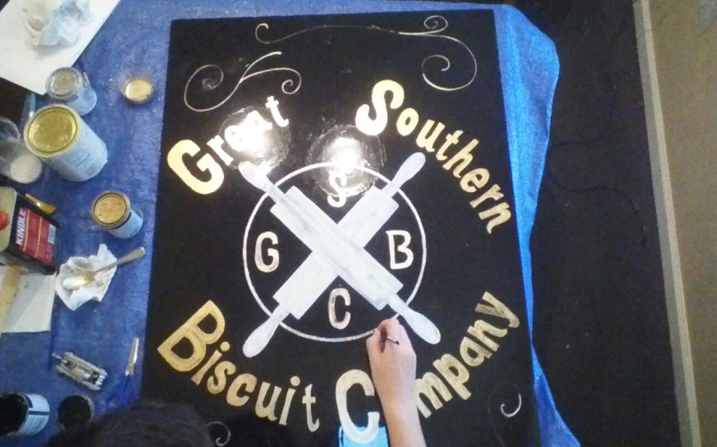 Enseigne noire du Great Southern Biscuit Company en cours de peinture, avec lettrage crème et ustensiles croisés.
