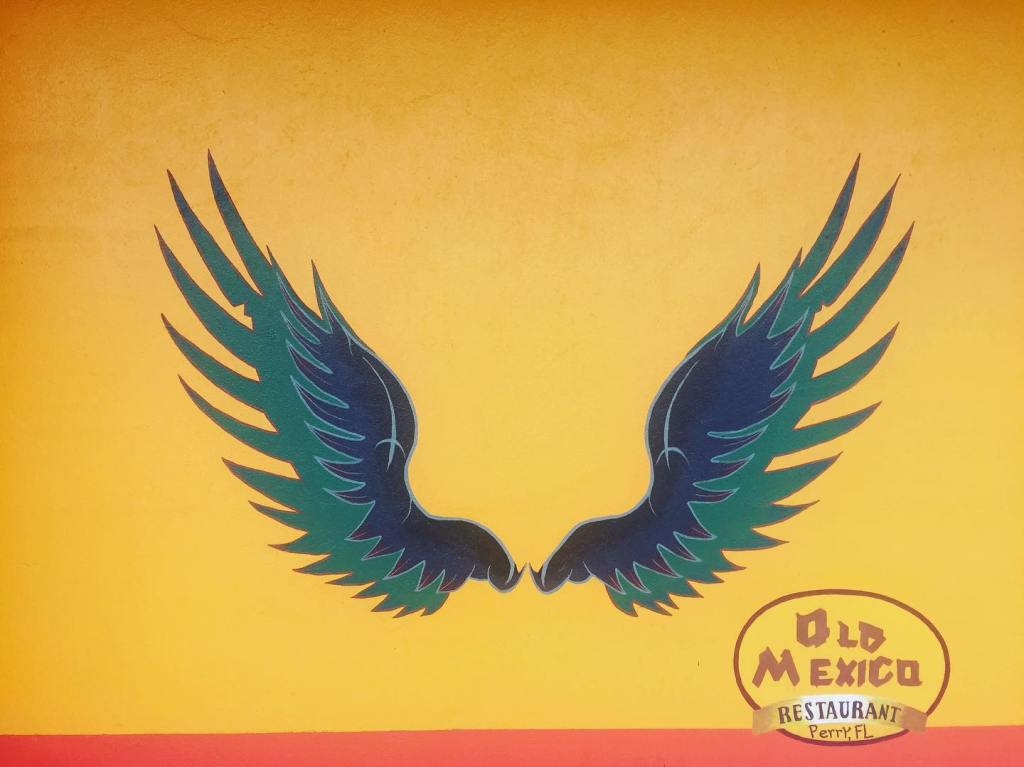 Deux grandes ailes bleu-vert peintes sur un mur jaune à côté du logo Old Mexico Restaurant.
