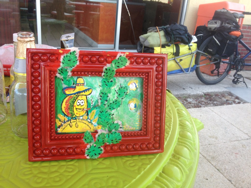 Petite peinture encadrée d’un cactus mexicain sur une table, avec un vélo de voyage chargé en arrière-plan.
