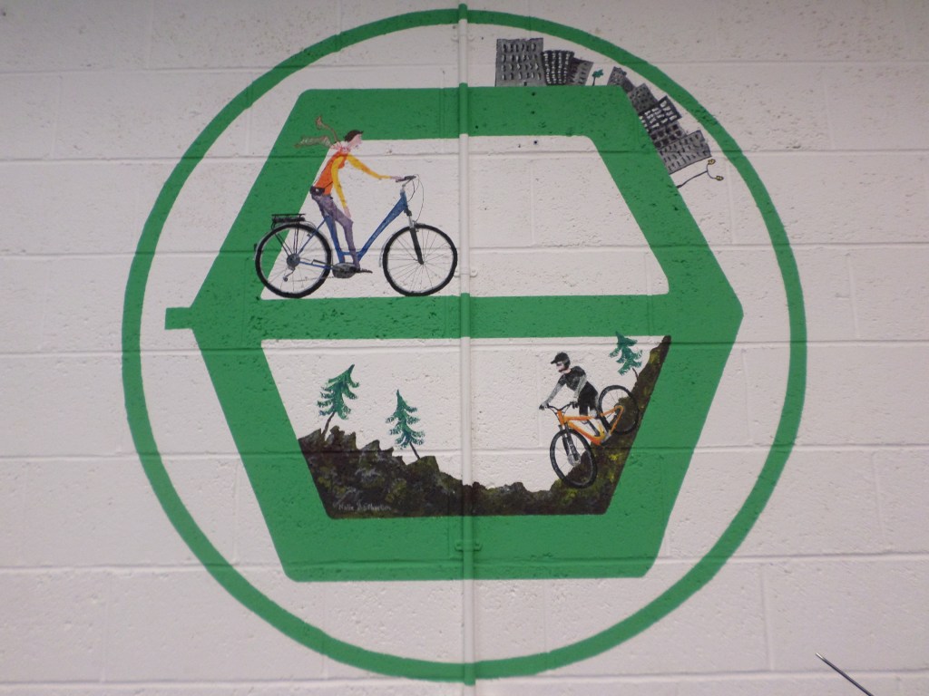 Fresque circulaire verte sur un mur blanc montrant deux cyclistes, des sapins et une route.
