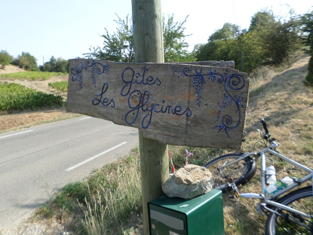 Ancienne enseigne en bois « Gîtes Les Glycines » peinte de motifs bleus et installée au bord d’une route.
