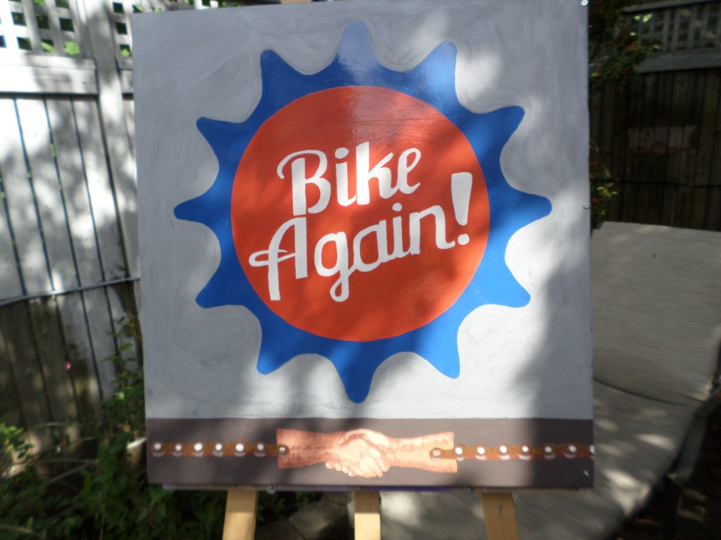Enseigne « Bike Again! » peinte en bleu, rouge et blanc, avec une poignée de mains au bas du panneau.
